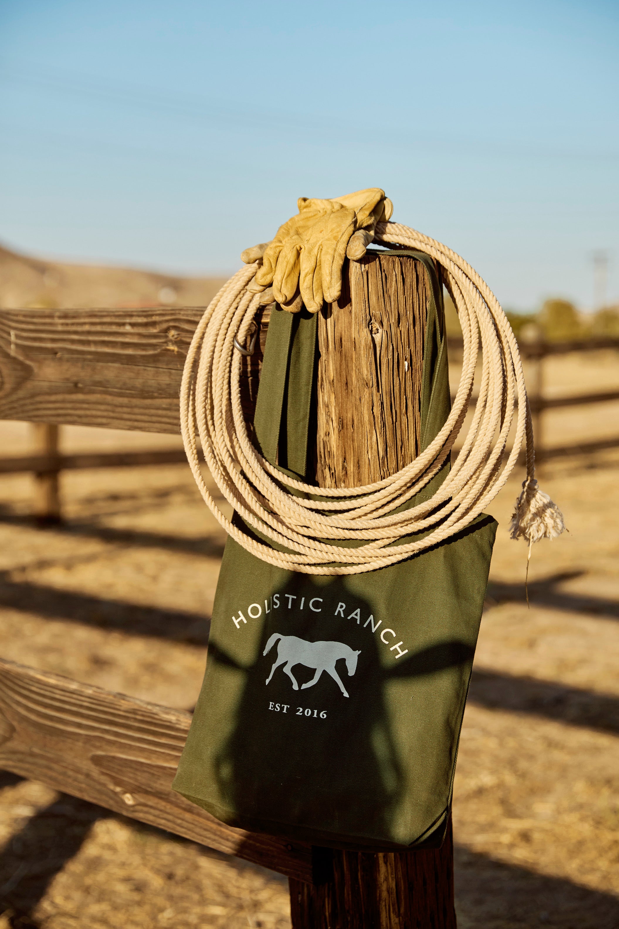 A **Holistic Ranch Tote** crafted from green organic certified cotton, featuring a horse logo and "Holistic Ranch, EST 2016" text, hangs on a wooden fence. A coiled rope and a pair of gloves rest on top, with a sunny ranch landscape in the background.